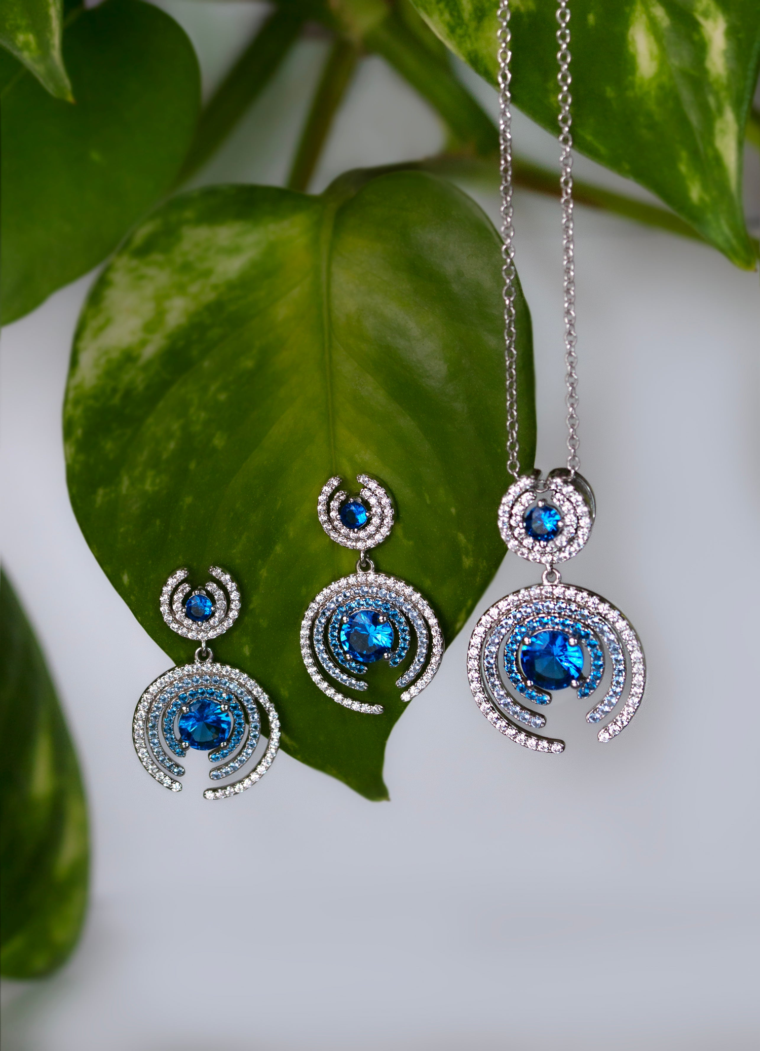 Exquisite jewelry set from the She is Nyxsara Collection featuring a platinum-plated necklace and matching earrings with vibrant blue sapphire-cut stones and spiral white zirconia design, displayed elegantly on fresh green leaves.