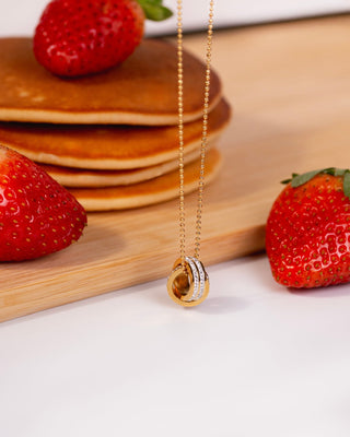 Gold necklace with a circular pendant on a wooden surface with strawberries and pancakes.