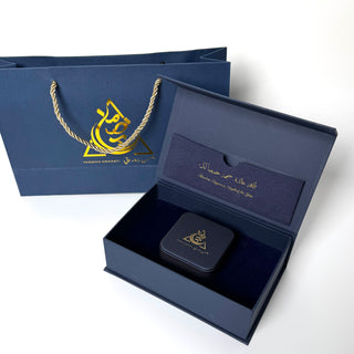 Open navy blue gift box with a black square item inside, accompanied by a matching navy blue bag with gold logo.