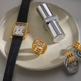 Gold watch with black strap, silver cylindrical object, and gold rings on a white dish.
