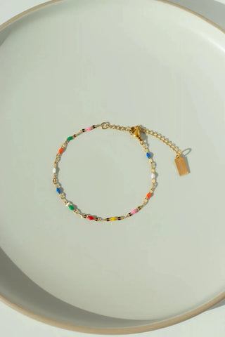 A delicate gold chain bracelet with small multicolored beads and a small rectangular tag, displayed on a light beige ceramic plate.