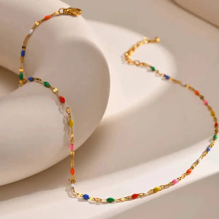 A gold chain necklace with small colorful gemstone-like beads in red, orange, yellow, green, blue, and pink, displayed on a white curved jewelry stand.