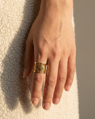Hand wearing a gold ring with a green gemstone on a textured beige surface