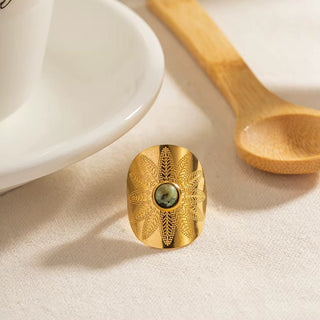 Gold ring with green stone on a beige surface with a wooden spoon and white dish in the background