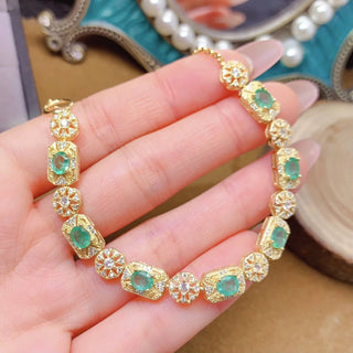 Gold bracelet with green natural zambian emerald gemstones held on a hand, with a blurred decorative background.
