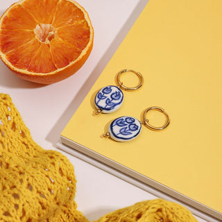 Pair of earrings with blue floral design on a yellow surface next to an orange and yellow fabric.